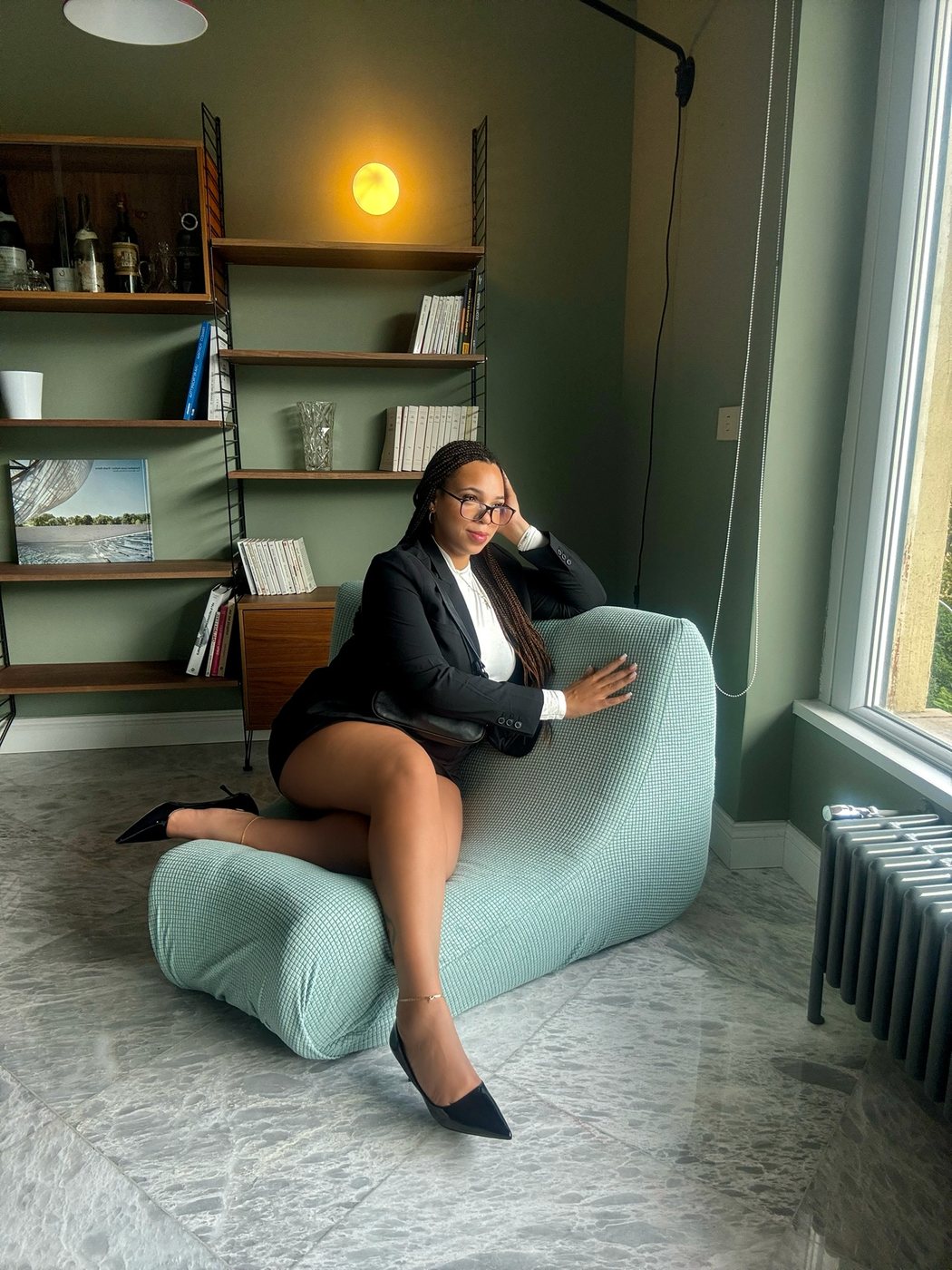 Delanna in a black blazer lounging on a pale green chair beside a window