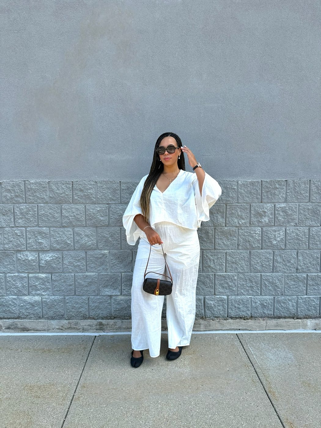 Delanna in an all-white set with sunglasses in front of a grey block wall